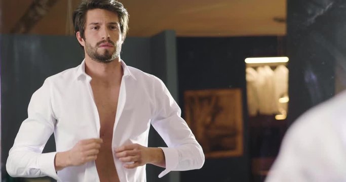 A handsome man in his loft dresses his sexy white shirt in a sexy way, looks into his room and smiles with a sure look at himself. Concept of: elegance, business, work.