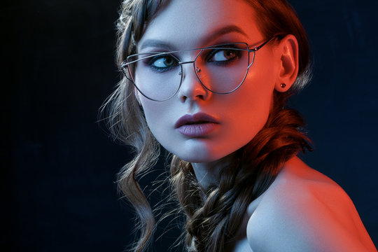 Close up studio portrait of beautiful woman with smoky eyes makeup wearing transparent glasses with metallic frame, posing in blue and red lights. Beauty, advertising concept. Copy, empty space 