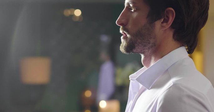 A handsome man in his loft dresses his sexy white shirt in a sexy way, looks into his room and smiles with a sure look at himself. Concept of: elegance, business, work.
