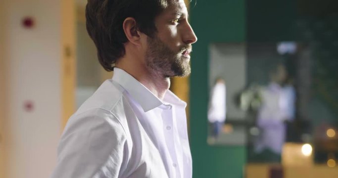 A handsome man in his loft dresses his sexy white shirt in a sexy way, looks into his room and smiles with a sure look at himself. Concept of: elegance, business, work.