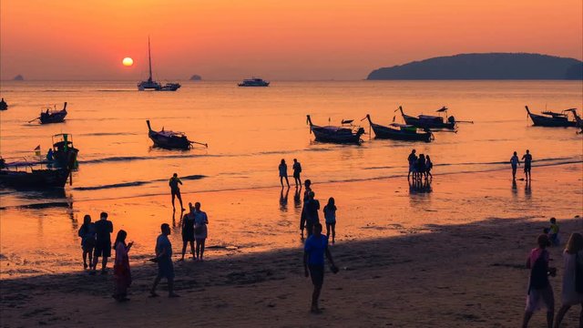 Time lapse of the Thai traditional beach with a lot of people silhouettes, moored Thai long-tail boats and yachts at sunset. Camera moves from left to right