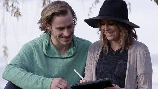 Panning up to young woman and man talking as they write on tablet while sitting at a park.