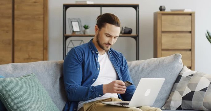 Young attractive man in jeans shirt sitting on the couch with a credit card in a hand and shopping online on the laptop computer in the cozy room. Inside