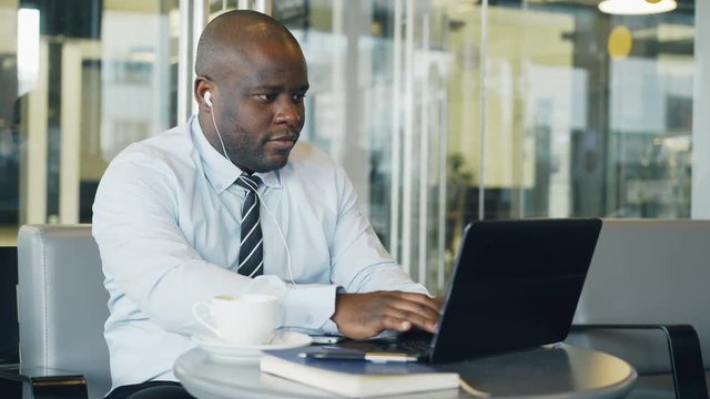 African American businessman in formal wear working and surfing social media on laptop while listening to music with earbuds in his ears in a glassy cafe