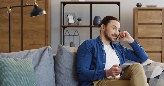 Caucasian young man in casual style sitting on the sofa with a tablet device in hands and talking on the phone. Indoor