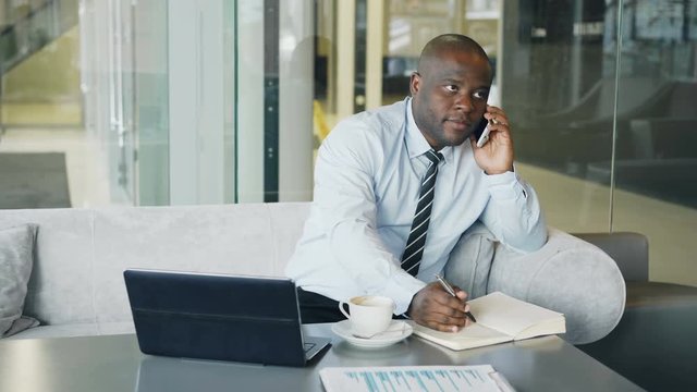 African American businessman talking on smartphone and writing down business information in his notepad sitting in glassy cafe with laptop and coffee on his table.
