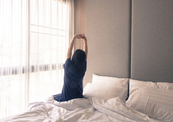 Fototapeta na wymiar Woman stretching in bed after waking up, back view. Woman sitting near the big white window while stretching on bed after waking up with sunrise at morning, back view.