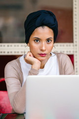 Portrait of a slim, young Muslim Malay woman sitting and working at a cafe during the day. She is...
