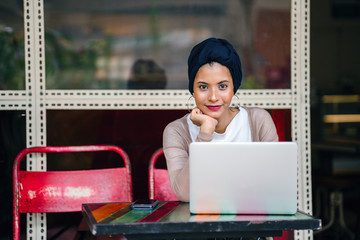 Portrait of a slim, young Muslim Malay woman sitting and working at a cafe during the day. She is...