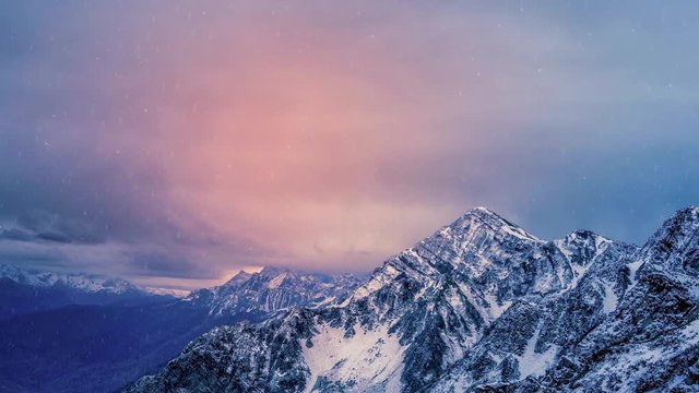 Light snow falling on the snowcapped mountains zooming in to reveal a stunning scene with a time lapse animated cloudscape during a dark sunrise or sunset