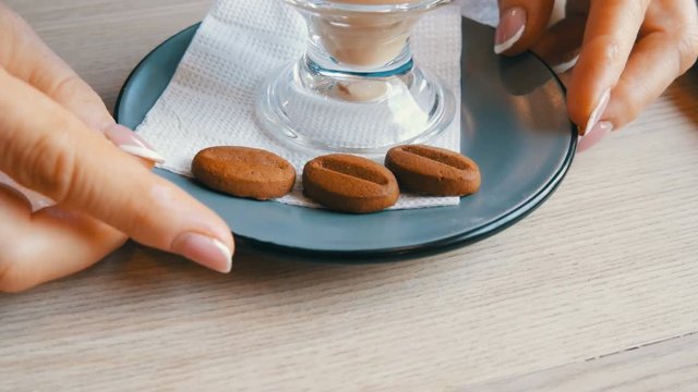 Woman is turning a saucer and a glass with a latte in a cafe, on the saucer lies a cookie in the form of coffee beans