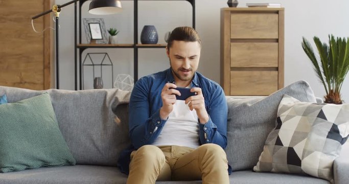 Excited caucasian man in the casual style sitting on the couch in the cozy room and playing games on the smartphone, then angry because loosing. At home. Inside