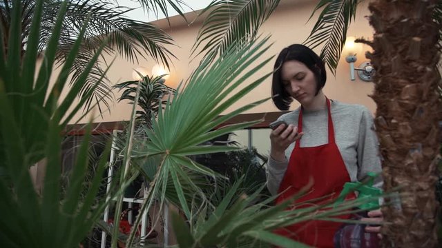 Young girl in red apron smiling using smartphone in greenhouse.