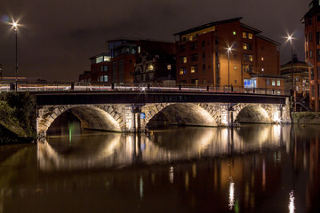 Night scene with lit bridge over a still river with long reflections in a residential area with low rise modern buildings.