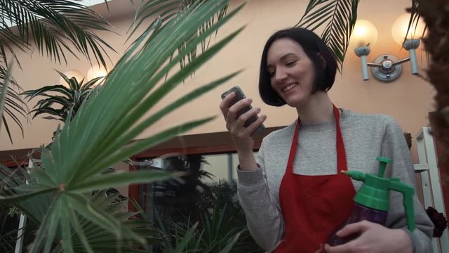 Young smily girl in red apron chatting online using smartphone in greenhouse