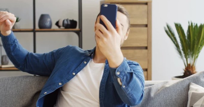 Young Caucasian man in the jeans shirt sitting on the sofa with pillows and having video chat on the smartphone. Inside