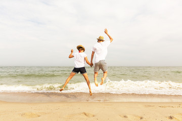 Two teenagers: a girl and a boy with blond hair, dressed in white T-shirts and shorts jumping up holding hands on a sea beach. Copy space.