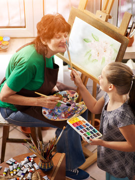 Artist painting easel in studio. Authentic grandmother and kids girl paints with palette watercolor paints palette and brush morning sunlight. Grandmother teaches grandsons of artist's profession.