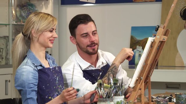 Handsome bearded man helping his beautiful girlfriend painting a picture working on a drawing at the Art Studio togetherness couple dating romance love affection artist creativity teamwork concept.