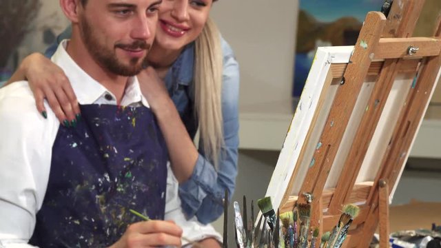 Sliding shot of a handsome tattooed bearded young male artist enjoying painting a picture at the Art Studio woth his beautiful happy girlfriend hugging him support happiness love dating romantic.