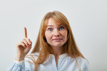 Headshot portrait of young european lady with red hair, white skin and freckles looking at the camera and showing at the information on left side of the picutre with her index finger raised.