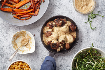 Flatlay with traditional swedish beef meatballs with gravy served in one portion black cast iron pan, handle wrapped in blue towel, plates with sauce, baked carrots and chickpeas. Dinner concept.