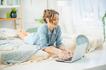 Young woman in blue shirt working with laptop lying on the bed in the bright and cozy bedroom at home