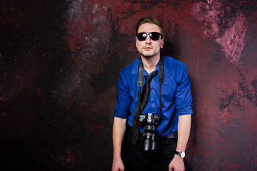 Studio portrait of stylish professional photographer man with camera, wear on blue shirt and necktie, sunglasses.