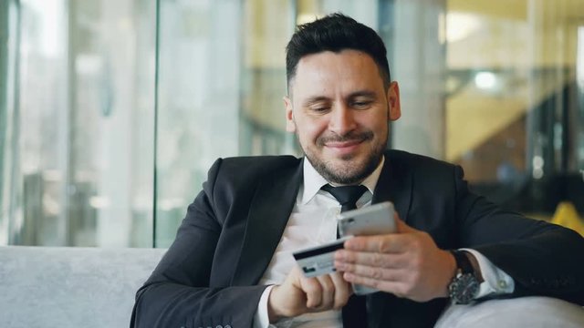 Smiling Caucasian businessman in suit using online banking holding credit card and smartphone in his hands in modern cafe during lunch break