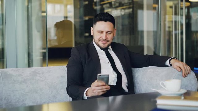 Happy Caucasian businessman in formal clothes smiling and looking at his smartphone screen while drinking coffee in airy cafe during lunch time