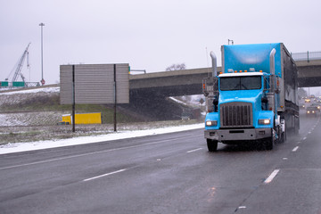 Fototapeta na wymiar Big rig blue classic semi truck with covered dry van semi trailer and high exhaust pipes is moving along multi-lane highway in snowy winter weather with poor visibility