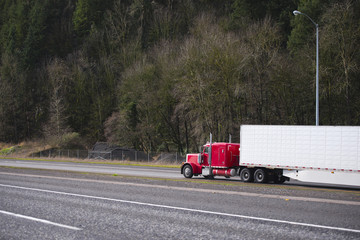 Big rig classic red semi truck with refrigerator semi trailer running on the wide road with trees on the background