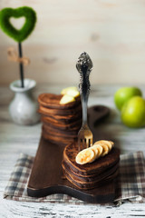 Obraz na płótnie Canvas Chocolate pancakes in the shape of heart with chopped banana slices on a textured wooden board. Pancakes close-up with a fork in them. In the background are green apples and a heart. Place for your te
