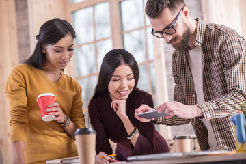 Modern technology. Pleasant nice bearded man holding his smartphone and taking a photo while standing together with his colleagues around the table