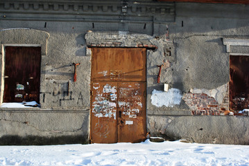Closed brown painted door with torn ads paper in which recognizable only separate letters, boarded up windows, gray plaster on wall, grunge background, winter snowy street, horizontal