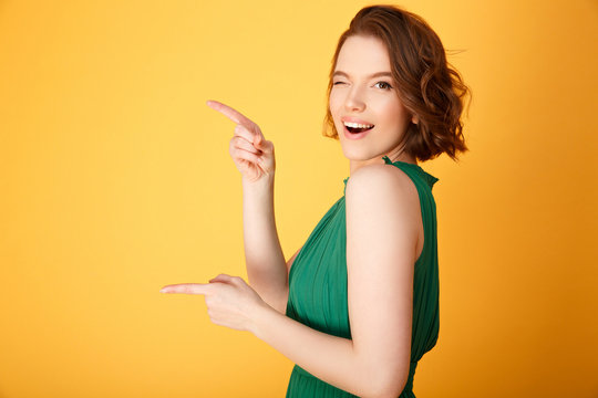 side view of attractive winking woman pointing away isolated on orange