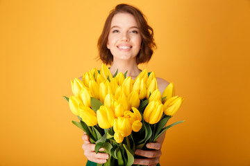 selective focus of smiling woman presenting bouquet of yellow tulips isolated on orange