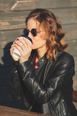 Young beautiful woman in round glasses and in a black leather jacket drinking coffee