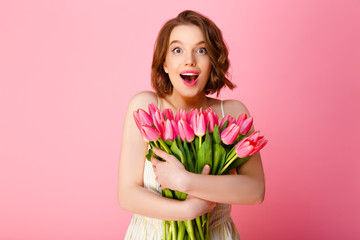 excited woman with bouquet of pink tulips looking at camera isolated on pink