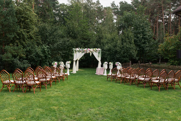 The beautiful platform for a wedding ceremony under the open sky: wooden chairs on a green grass, the square arch decorated with fresh flowers