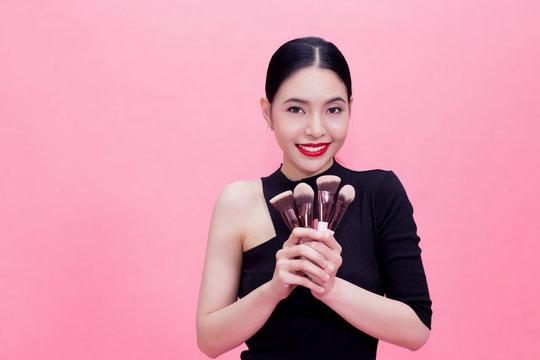 Young Stylish Asian woman carrying makeup brushes on hand with copy space. Beautiful fashionable woman isolated over pink background.