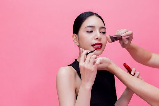 Beautiful and luxurious young busy Asian woman doing make-up with extra helping hands to help isolated over pink background.