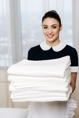 young happy maid in uniform holding stack of clean towels and looking at camera