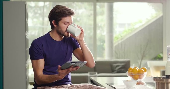 A man in the kitchen while having breakfast and sipping tea or milk in the cup sends a message or calls with the tablet and smiles. Concept of: social network, message, technology.