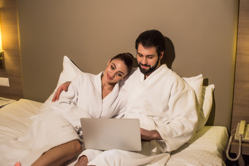 happy couple in bathrobes using laptop together in bed of hotel suite