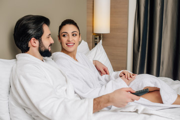 Obraz premium happy couple in bathrobes with tv remote control relaxing in bed at hotel suite