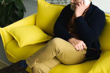 cropped shot of senior man holding eyeglasses and talking by smartphone while sitting on sofa