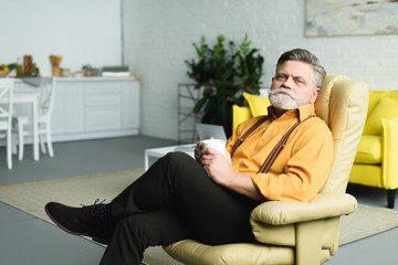 handsome bearded senior man holding cup of tea and looking at camera while sitting on armchair at home