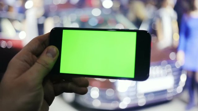 man hand holding smart phone mobile in front of a new car / close-up blur expensive luxury lifestyle chroma key green sreen horizontal keeping photo taking picture auto show showroom notification
