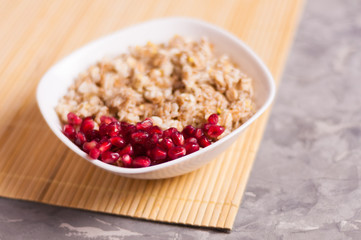 Cooked yummy warm oatmeal and heap of ripe fresh pomegranate seeds in white ceramic bowl on clean bamboo rug on worn gray scratched concrete